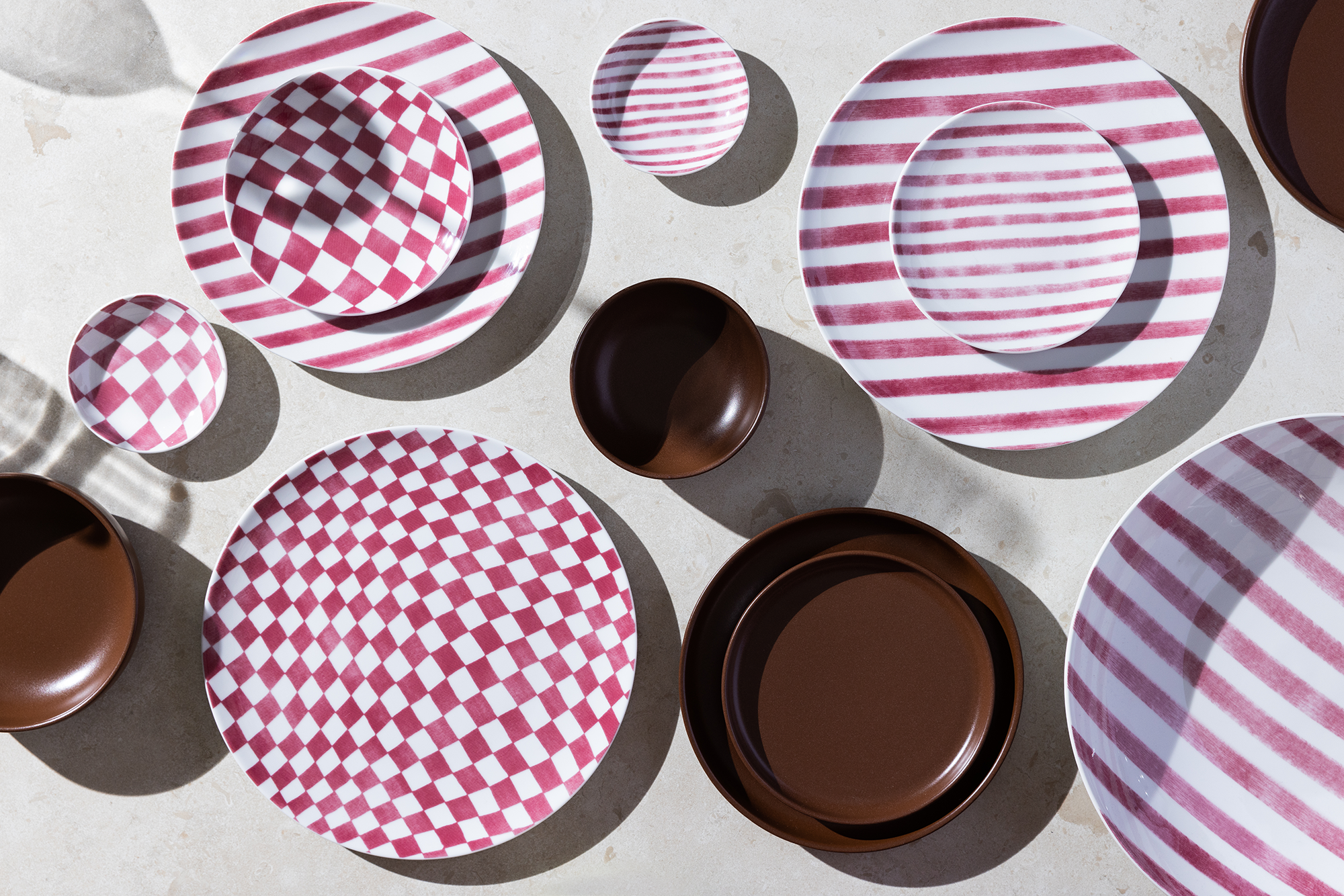 Vinröd och vit randiga och rutiga tallrikar i porslin tillsammans med mörkt bruna tallrikar i brunt stengods. Uppdukade på ett ljust bord
