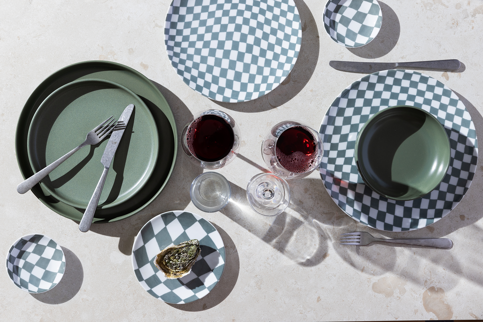 Vit och grön rutiga porslinstallrikar tillsammans med gröna tallrikar i stengods på ett ljust bord. 2 vinglas upphällda, ett par bestick och ett ostron har använts som props.