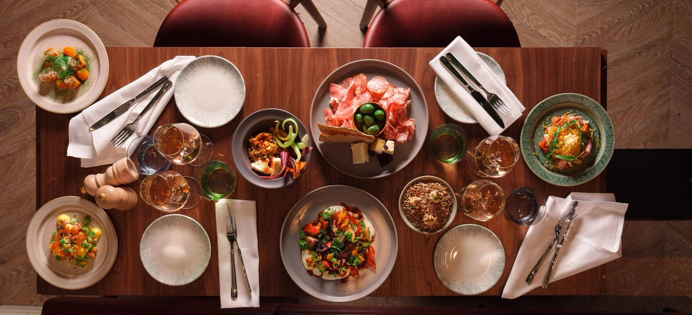 Dukat bord med en mix av tallrikar i olika storlekar. Bordet är av brunt trä och tallrikarna går i grått, beige och grönt. Maten ser inbjudande ut och det finns 4 servetter och bestick.