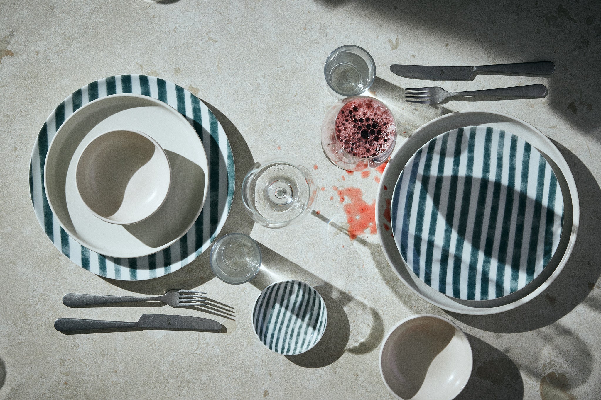 Randiga tallrikar i porslin tillsammans med vita tallrikar i stengods uppdukade på ett ljust bord. Ett glas rödvin finns upphällt och vinfläckar ger dynamik åt bilden