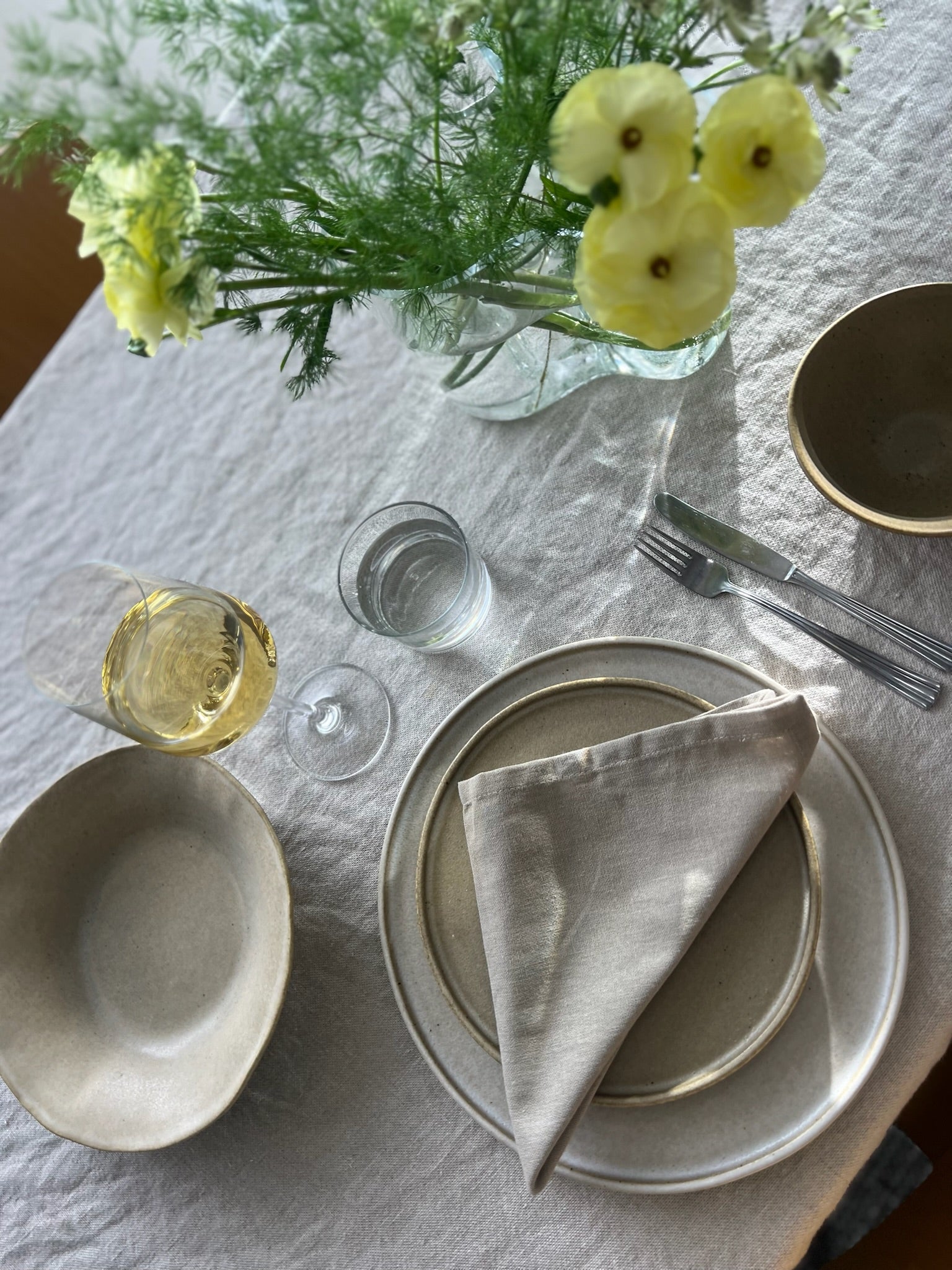 Dukat bord med en ljus linneduk. Tallrikar i stengods i Sand och mörk beige. Vacker gul blomma och ett glas vitt vin upphällt