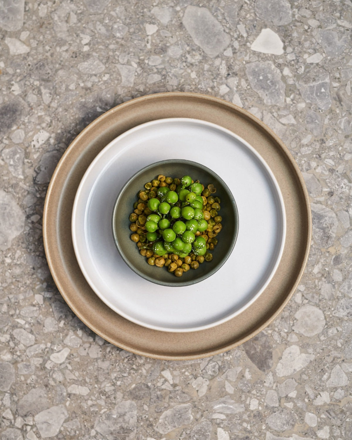 Sandfärgad stor tallrik med en mindre vit glansig stengodstallrik ovanpå, samt en mindre grön skå med ärtor